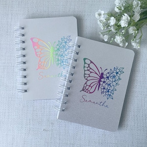 May include: Two spiral-bound notebooks with butterfly and floral designs. The butterflies are iridescent with rainbow colours. The name "Samantha" is printed below the butterfly. The notebooks are white and silver.