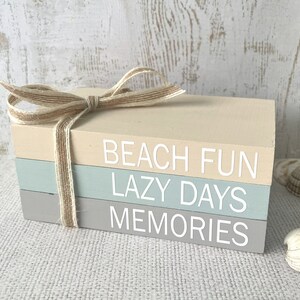 May include: A stack of three wooden blocks with the words "Beach Fun", "Lazy Days", and "Memories" painted in white. The blocks are tied together with a brown ribbon and are sitting on a beige surface.