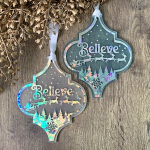 Op de afbeelding: Twee iriserende kerstversieringen met een vierpasvorm. Het woord "Believe" is cursief geschreven. De ornamenten tonen een door rendieren getrokken slee, bomen en sneeuwvlokken. Elk ornament is ongeveer 10 cm hoog en heeft een wit lint om op te hangen.