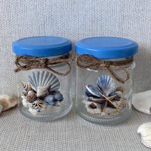 May include: Two clear glass jars with bright blue lids, each tied with a rustic brown twine bow. Inside each jar is a decorative arrangement of various seashells and a small starfish resting on a sandy base, creating a coastal display.