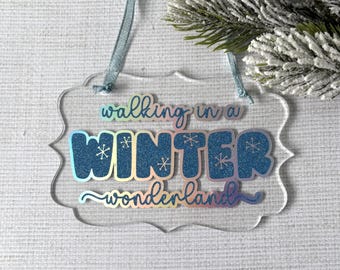 May include: Clear acrylic Christmas ornament with the words "walking in a WINTER wonderland" in blue glitter and iridescent lettering. The ornament has a scalloped edge and a light blue ribbon. A sprig of faux evergreen is in the background.