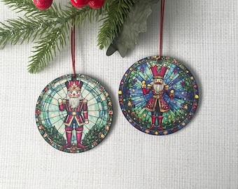 May include: Two round Christmas ornaments featuring a nutcracker design. The ornaments have a stained-glass effect with a red, blue, and green color scheme. Each ornament is suspended by a red ribbon, and a sprig of greenery with red berries is in the background.