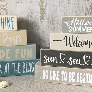 May include: A stack of five wooden blocks with painted text, each block is a different colour. The top block is grey and says "Hello Summer!" The next block is white and says "Welcome". The next block is grey and says "sun sea sand". The next block is white and says "I do like to be beside the seaside". The bottom block is blue and says "Life is better at the beach".