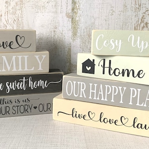 May include: Stacked wooden blocks with a home theme. The blocks are painted in shades of grey, black, and cream. The top block reads "love" with a heart symbol. The next block reads "FAMILY". The third block reads "home sweet home". The fourth block reads "this is us OURLIFE OUR STORY OUR". The second set of blocks reads "Cosy Up" on the top block, "Home" with a house and heart symbol on the second block, "OUR HAPPY PLACE" on the third block, and "live love laugh" on the bottom block.