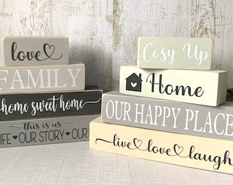 May include: Stacked wooden blocks with a home theme. The blocks are painted in shades of gray, black, and cream. The top block reads "love" with a heart symbol. The next block reads "FAMILY". The third block reads "home sweet home". The fourth block reads "this is us OURLIFE OUR STORY OUR". The second set of blocks reads "Cosy Up" on the top block, "Home" with a house and heart symbol on the second block, "OUR HAPPY PLACE" on the third block, and "live love laugh" on the bottom block.