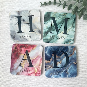 May include: Four square coasters with a marble pattern in different colours. Each coaster has a letter and a name printed on it. The letters are H, M, A, and D. The names are Harry, Michelle, Abbie, and Darren.