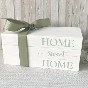 Op de afbeelding: Witte decoratieve houten blokken gestapeld met de woorden "HOME sweet HOME" in groene letters. Een saliegroen lint en strik met een decoratieve sluiting wikkelen de blokken. Een woondecoratie-artikel.