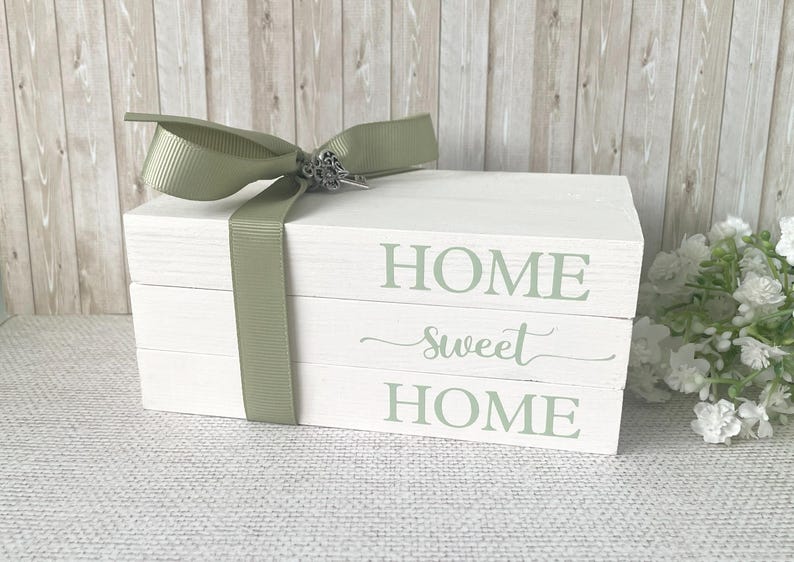 Op de afbeelding: Witte decoratieve houten blokken gestapeld met de woorden "HOME sweet HOME" in groene letters. Een saliegroen lint en strik met een decoratieve sluiting wikkelen de blokken. Een woondecoratie-artikel.
