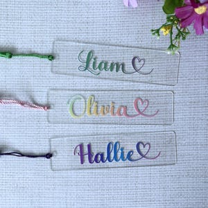 Op de afbeelding: Drie transparante acryl bladwijzers met kleurrijke tekst en kwastjes. De namen "Liam", "Olivia" en "Hallie" zijn in script geschreven met een hartsymbool. Elke bladwijzer heeft een kwastje in een andere kleur: groen, roze en paars.