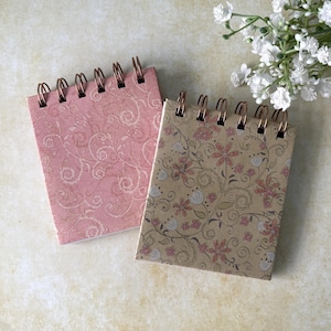 May include: Two small spiral-bound notebooks with floral patterns. One has a pink cover with a swirling design, the other a tan cover with pink and white flowers. The spiral binding is copper-coloured.