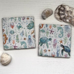 May include: Two square coasters featuring a watercolour print of marine life including sea turtles, seahorses, jellyfish, crabs, starfish, and coral on a light background, accompanied by several natural seashells of varying shapes and sizes.