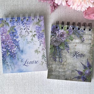 May include: Two spiral-bound notebooks. One has a watercolour design of purple wisteria and the name "Louise". The other has a floral design with sheet music and the name "Katie". Both have a soft, vintage aesthetic.