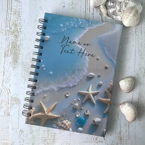 May include: A spiral bound notebook with a beach scene on the cover. The cover features a blue ocean, white sand, and seashells. The text "Name or Text Here" is printed on the cover.
