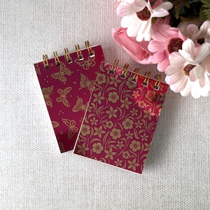 May include: Two spiral-bound notebooks with a deep magenta background. One notebook features a gold-toned butterfly pattern, while the other displays a gold-toned floral and vine design. Both notebooks have gold-coloured metal wire binding at the top.