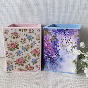 May include: Two decorative wooden pen holders. One is pink and covered in a floral pattern of pink and blue roses. The other is blue and features a watercolor painting of purple wisteria.