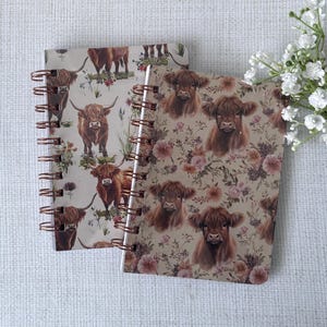 May include: Two spiral-bound notebooks with Highland cow illustrations. One notebook features full-body cow images with floral accents, while the other showcases close-up cow faces amidst a floral pattern. The notebooks have copper-colored spiral bindings.