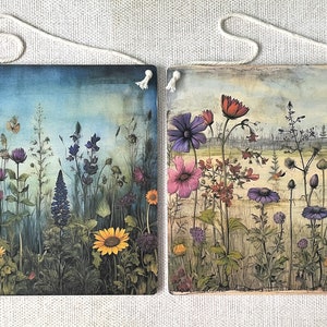 Op de afbeelding: Twee vierkante houten wanddecoraties met een bloemenmotief. Het linker paneel heeft een blauwe en groene achtergrond met een verscheidenheid aan bloemen in tinten van paars, roze, geel en oranje. Het rechter paneel heeft een beige achtergrond met een verscheidenheid aan bloemen in tinten van paars, roze en rood.