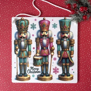 May include: A square Christmas decoration featuring three colourful nutcracker soldiers. The nutcrackers are in teal, pink, and gold with white fur trim. The decoration has the words "Merry Christmas" on it.