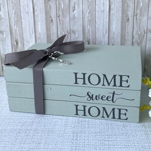 Op de afbeelding: Een stapel van drie houten blokken, geverfd in een gedempte groen-grijze kleur. De blokken zijn samengebonden met een grijs lint en voorzien van de tekst "HOME sweet HOME" in zwarte letters. Een zilveren bedel hangt aan het lint.