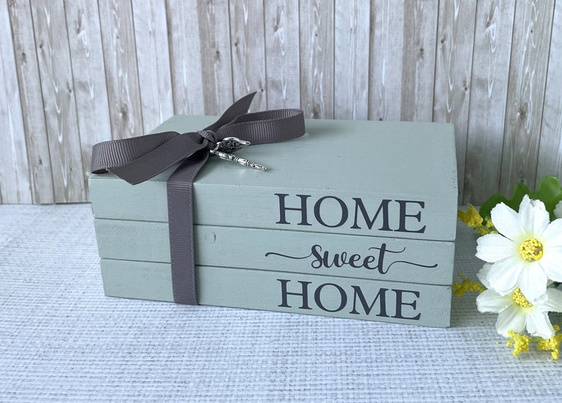 Op de afbeelding: Een stapel van drie houten blokken, geverfd in een gedempte groen-grijze kleur. De blokken zijn samengebonden met een grijs lint en voorzien van de tekst "HOME sweet HOME" in zwarte letters. Een zilveren bedel hangt aan het lint.