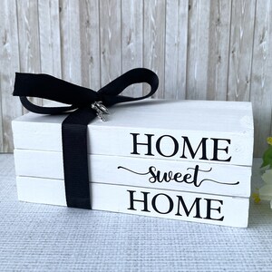 Op de afbeelding: Witte houten blokdecoratie met de woorden "HOME sweet HOME" in zwarte letters. Een zwart lint is om de blokken gewikkeld en vastgeknoopt met een strik. Een kleine zilveren bedel is aan de strik bevestigd.