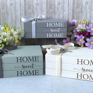 May include: Three decorative wooden book stacks in gray, sage green, and white, each with the words "HOME sweet HOME" printed on them. The stacks are tied with ribbons and adorned with decorative keys. Floral arrangements in yellow and purple are in the background.