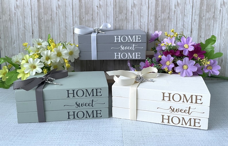 Op de afbeelding: Drie decoratieve houten boekenstapels in grijs, saliegroen en wit, elk met de woorden "HOME sweet HOME" erop gedrukt. De stapels zijn vastgebonden met linten en versierd met decoratieve sleutels. Bloemschikkingen in geel en paars staan op de achtergrond.