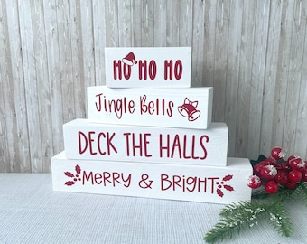 May include: Four white wooden blocks stacked to create a Christmas-themed display. Each block features red text: "HO HO HO", "Jingle Bells", "Deck the Halls", and "Merry & Bright". Festive holiday decor.