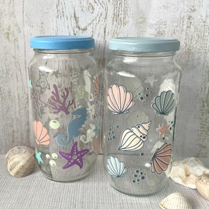 May include: Two glass jars with blue lids decorated with sea-themed designs. The jars are filled with various sea creatures, shells, and coral. The jars are on a light brown surface.