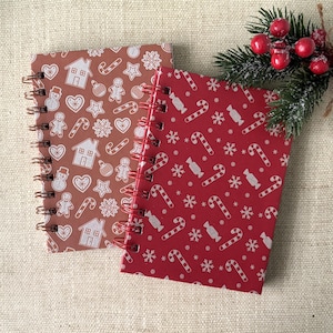 Op de afbeelding: Twee spiraalgebonden notitieboekjes met feestelijke kerstpatronen. Eén notitieboekje is rood met witte zuurstokken en sneeuwvlokken. De andere is bruin met peperkoekmannetjes, huisjes en sterren. Een takje groen met rode bessen ligt in de buurt.