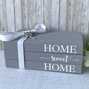 Op de afbeelding: Grijze decoratieve boekenstapel met de woorden "HOME sweet HOME" in witte letters. Een wit lint met een sleutelbedel is om de boeken gewikkeld. Het is een woondecoratie-item.