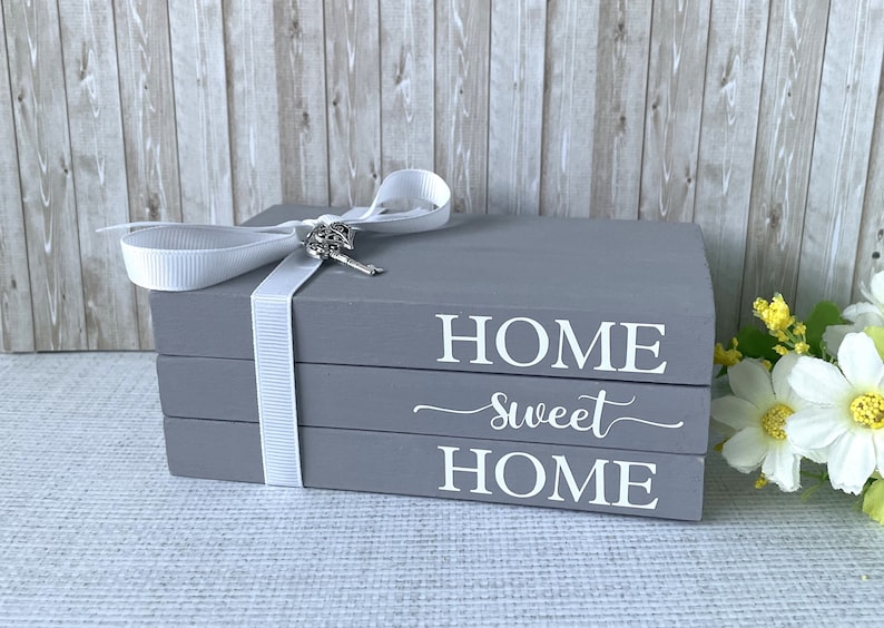 Op de afbeelding: Grijze decoratieve boekenstapel met de woorden "HOME sweet HOME" in witte letters. Een wit lint met een sleutelbedel is om de boeken gewikkeld. Het is een woondecoratie-item.