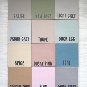 Op de afbeelding: Een verzameling van twaalf vierkante houten tegels, elk geschilderd in een andere kleur. De kleuren zijn onder meer Greige, New Sage, Light Grey, Urban Grey, Taupe, Duck Egg, Beige, Dusky Pink, Teal, Golden Sand, Pink en Dark Grey. Elke tegel heeft een kleurnaam in zwarte tekst.