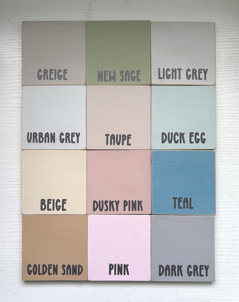 Op de afbeelding: Een verzameling van twaalf vierkante houten tegels, elk geschilderd in een andere kleur. De kleuren zijn onder meer Greige, New Sage, Light Grey, Urban Grey, Taupe, Duck Egg, Beige, Dusky Pink, Teal, Golden Sand, Pink en Dark Grey. Elke tegel heeft een kleurnaam in zwarte tekst.