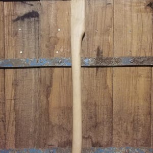 Könnte beinhalten: Ein neuer, unfertiger Axtstiel aus hellfarbenem Holz. Der Stiel ist lang und schlank, mit einem gebogenen Griff und einer flachen Oberseite zur Befestigung des Axtkopfes. Der Hintergrund ist eine Holzoberfläche.