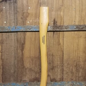 Könnte beinhalten: Ein hellfarbener Holzbeilgriff mit geschwungenem Design. Der Text "SMEDBERG HICKORY #LAC01" ist vertikal auf dem Griff aufgedruckt. Der Hintergrund ist eine Holzoberfläche mit Metallakzenten.