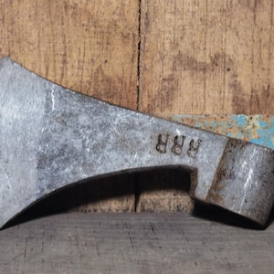 May include: A weathered metal axe head with a curved blade and a cylindrical eye for the handle. The axe head has the letters "RRR" stamped on it. The background is a rustic wooden surface.