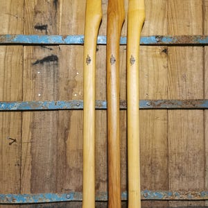 Puede incluir: Tres mangos de hacha de madera en tonos marrones y beige se muestran contra un fondo de madera rústica. Cada mango tiene un diseño curvo y un agujero en la base para asegurar la cabeza del hacha. Los mangos son de diferentes tipos de madera.