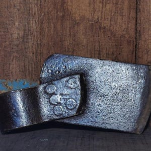 Può includere: Primo piano di due teste d'ascia vintage. La testa più grande ha una superficie argentata strutturata. La testa più piccola è grigio scuro con rivetti. Lo sfondo è una superficie di legno usurata.