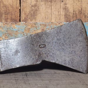 Op de afbeelding: Een verweerde metalen bijlkop met een rechthoekige vorm en een licht gebogen blad. De bijlkop heeft een versleten, getextureerd oppervlak met een kleine, rechthoekige markering. De achtergrond toont verweerd hout en roestig, blauw geschilderd metaal.