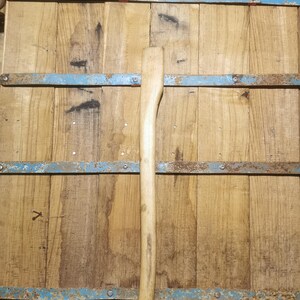 Pode incluir: Um cabo de machado de madeira clara contra um fundo de tábuas de madeira desgastadas. O cabo é longo e fino, com uma extremidade curva para agarrar. As tábuas são presas por faixas de metal enferrujado com tinta azul.
