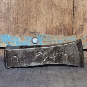 Può includere: Una testa d'ascia in metallo usurato con segni visibili. La testa dell'ascia è di colore metallico scuro, che mostra segni di usura. Lo sfondo presenta una superficie in legno e una striscia di metallo blu.