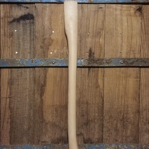 Könnte beinhalten: Ein neuer, unfertiger Axtstiel aus hellfarbenem Holz. Der Stiel ist lang und schlank, mit einem gebogenen Ende zum Greifen. Der Hintergrund ist eine Holzoberfläche mit blauen Metallakzenten.