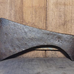 Könnte beinhalten: Ein verwitterter, antiker Axtkopf mit gebogener Klinge und dunkler, strukturierter Oberfläche. Der Axtkopf besteht aus Metall und weist Gebrauchsspuren auf. Der Axtkopf wird vor einem Holzgrund gezeigt. Ein historisches Werkzeug.