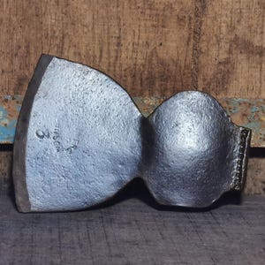 May include: A vintage axe head with a silver finish. The axe head is marked with the number "2" and the letters "F.R.C." on the side. The axe head is set against a rustic wooden background.