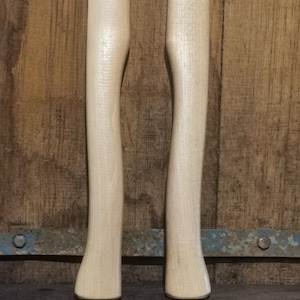 Op de afbeelding: Twee onafgewerkte houten bijlhandvatten, naast elkaar, tegen een rustieke houten achtergrond. De handvatten zijn licht van kleur, met een glad, gebogen ontwerp. De achtergrond is donker, verweerd hout.