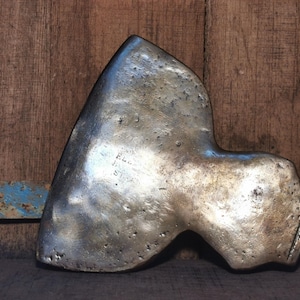 Pode incluir: Uma cabeça de machado metálica desgastada com uma superfície texturizada. A cabeça do machado tem um topo triangular e uma seção inferior curva. O metal tem uma cor prateada opaca com algumas manchas mais escuras. O fundo é uma superfície de madeira.