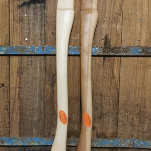 Pode incluir: Duas cabos de machado de madeira, um de cor clara e outro castanho escuro. Cada cabo tem um autocolante oval laranja. Os cabos são exibidos contra um fundo de madeira rústica com detalhes metálicos azuis.