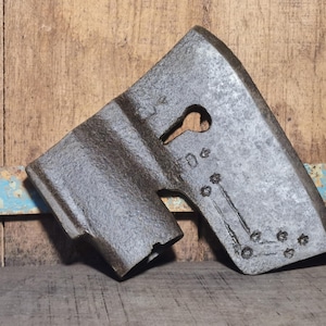 Op de afbeelding: Een verweerd, grijs metalen object met een sleutelgatvormige opening en geëtste markeringen. Het object heeft een ruw, getextureerd oppervlak en lijkt een onderdeel van een industrieel gereedschap te zijn. De achtergrond is een rustiek houten oppervlak.