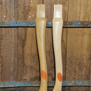 Op de afbeelding: Twee houten bijlhandvatten met gebogen schachten. Eén handvat is lichtbruin, de andere is van nature houtkleurig. Beide hebben oranje en witte labels bij de basis. De achtergrond is een rustieke houten muur. De handvatten zijn ongeveer 40 cm lang.